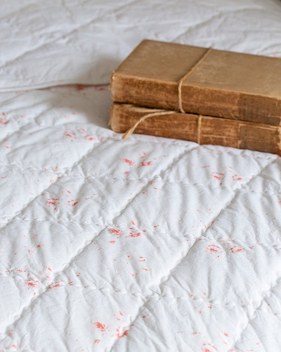 A close-up of a white quilted bedspread with small red speckles, with two old brown books tied with string resting on the top right corner.