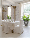 A bright, elegant dining room with a long table set for six in white linens, surrounded by white upholstered chairs. The room features lavender and white floral wallpaper, tall windows with white curtains, and a large potted green plant in a galvanized bucket on the floor.