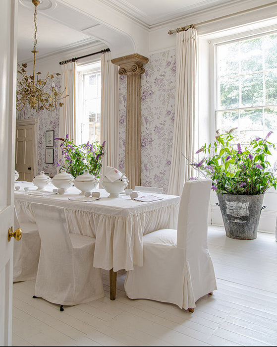 A bright, elegant dining room with a long table set for six in white linens, surrounded by white upholstered chairs. The room features lavender and white floral wallpaper, tall windows with white curtains, and a large potted green plant in a galvanized bucket on the floor.