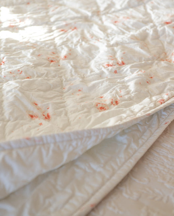 A close-up of a white quilted mattress protector or comforter with scattered small red stains, shown on a bed.